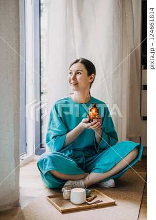 Young woman sitting on the floor holding a lit amber glass candle beside a tray with incense and tea. Spiritual minimalism at home, quiet luxury, cozy meditation, sacred simplicity 124661814