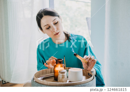 Young woman burning palo santo near a tray with candles and tea in a calm home setting. Sensory mindfulness for daily balance, palo santo ritual, inner calm, grounding therapy 124661815