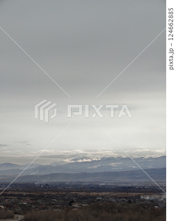 Overcast sky blankets a distant mountain range with a town nestled in the valley. A moody and expansive scene portraying the quiet of nature and settlement below. 124662885