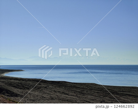 A scenic view of a tranquil coastline with calm blue waters and a faint outline of snow-capped mountains in the distance under a bright sky. 124662892