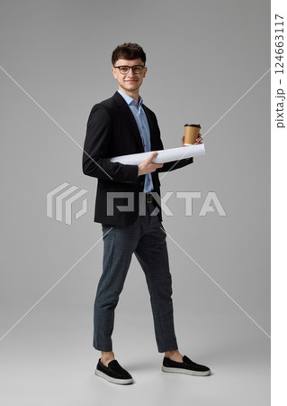 Full length portrait of confident young businessman in suit holding coffee and blueprint against grey background. Concept of ambition Full length portrait of confident young businessman in suit holding coffee and blueprint against grey background. Concept of ambition 124663117
