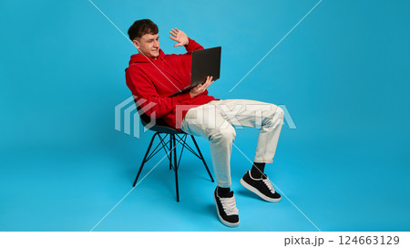 Relaxed young man on chair waving to laptop screen during online conversation, against blue background. Concept of distance work Relaxed young man on chair waving to laptop screen during online conversation, against blue background. Concept of distance work 124663129
