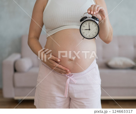 Close up of pregnant woman's belly holding alarm clock. 124663317