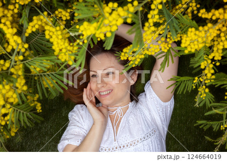 Happy woman in a white dress lying on the grass, surrounded by bright yellow mimosa branches, enjoying the fresh spring atmosphere. A moment of joy, nature, and relaxation. Happy woman in a white dress lying on the grass, surrounded by bright yellow mimosa branches, enjoying the fresh spring atmosphere. A moment of joy, nature, and relaxation. 124664019