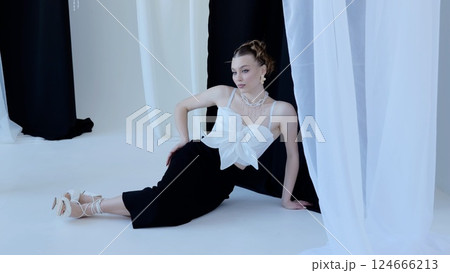 Fashion model wearing a white top and black pants is posing on the floor in a studio with black and white curtains, creating a contrasting and stylish atmosphere Fashion model wearing a white top and black pants is posing on the floor in a studio with black and white curtains, creating a contrasting and stylish atmosphere 124666213