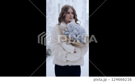 Stylish woman in white fur coat gripping frosted evergreen branches, standing amid snow covered urban landscape during chilly winter season 124666216