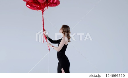 Fashion model posing in sleek black dress, holding vivid red heart balloons against white backdrop, radiating elegant romance and festive love atmosphere Fashion model posing in sleek black dress, holding vivid red heart balloons against white backdrop, radiating elegant romance and festive love atmosphere 124666220