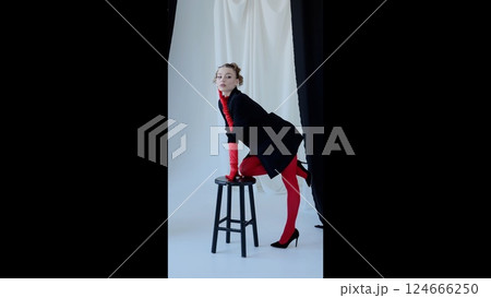 Confident female model posing elegantly in vibrant red gloves and tights, leaning on stool with delicate hand touching face against monochrome studio backdrop Confident female model posing elegantly in vibrant red gloves and tights, leaning on stool with delicate hand touching face against monochrome studio backdrop 124666250