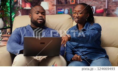 African american employees in living room using laptop and preparing presentation, inserting charts and adjusting layouts. Young couple finishing company project and writing summaries. Camera B. African american employees in living room using laptop and preparing presentation, inserting charts and adjusting layouts. Young couple finishing company project and writing summaries. Camera B. 124669493