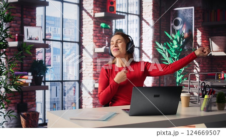 Mature person having fun solving her work tasks with headset on at desk, workflow and productivity with focus music in modern loft. Woman freelancer with lovely vibes working remotely. Camera B. Mature person having fun solving her work tasks with headset on at desk, workflow and productivity with focus music in modern loft. Woman freelancer with lovely vibes working remotely. Camera B. 124669507