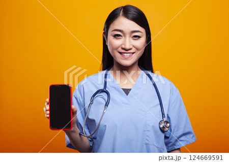 Young doctor in medical uniform, holding smartphone with black screen against a yellow background, gazing at camera. Portrait of asian woman in nurse scrubs, grasping mobile device with blank display. 124669591