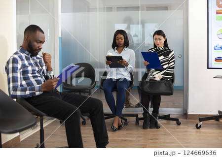 Small group of applicants in office lobby, reading company internal regulations and preparing for employment meeting. Multiethnic unemployed people sitting in queue for job interview process. 124669636