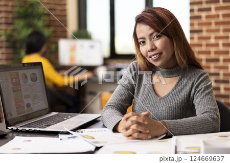 Asian businesswoman sits confidently at her office desk, smiling at the camera, ready to tackle projects. Portrait of female professional prepared for busy workday, surrounded by paperwork and laptop. Asian businesswoman sits confidently at her office desk, smiling at the camera, ready to tackle projects. Portrait of female professional prepared for busy workday, surrounded by paperwork and laptop. 124669637
