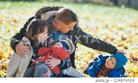 Family cuddling newborn, caressing toddler among fallen leaves during golden autumn sunlight in peaceful park setting Family cuddling newborn, caressing toddler among fallen leaves during golden autumn sunlight in peaceful park setting 124674324