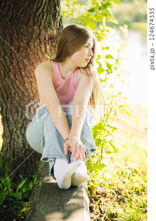 Serene female resting near lakeside tree, basking in warm sunlight, peaceful expression reflecting calm natural surroundings 124674335