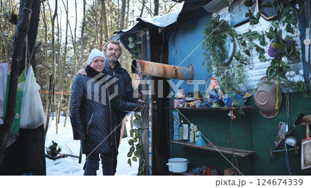 Homeless couple standing beside ramshackle shelter, weathering winter landscape with quiet determination amid challenging circumstances Homeless couple standing beside ramshackle shelter, weathering winter landscape with quiet determination amid challenging circumstances 124674339