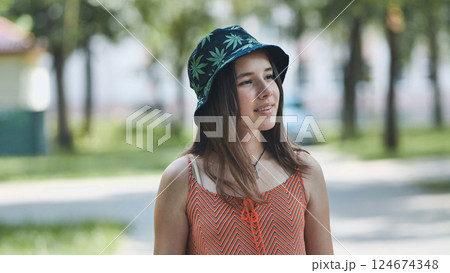 Teenage girl grinning, sporting marijuana leaf bucket hat while relaxing outdoors during sunny day in green park setting Teenage girl grinning, sporting marijuana leaf bucket hat while relaxing outdoors during sunny day in green park setting 124674348