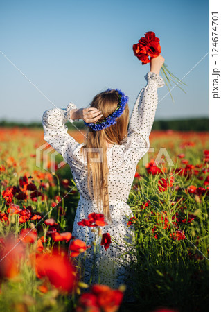 Blonde woman in white polka dot dress, flower crown, holding red poppies amid sunny wildflower meadow Blonde woman in white polka dot dress, flower crown, holding red poppies amid sunny wildflower meadow 124674701