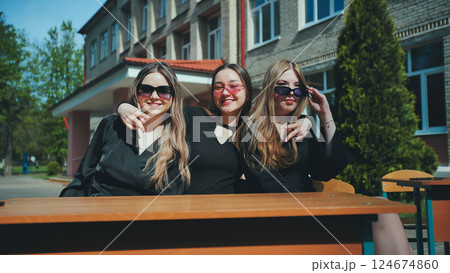 Three cheerful students in sunglasses sit at an outdoor school desk, smiling on a sunny day 124674860