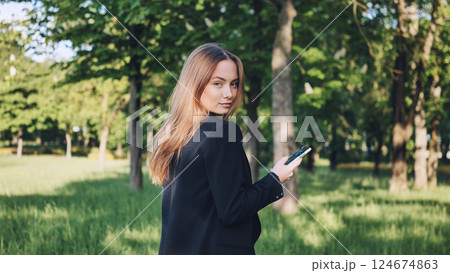 Young businesswoman holding mobile phone walking in green city park looking back over shoulder 124674863