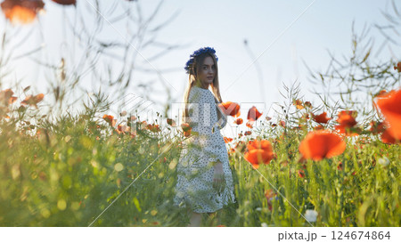 Young woman with long hair and flower crown walking in a poppy field during a sunny summer day 124674864