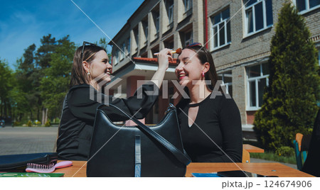 Two cheerful university students enjoying their break, applying makeup together on a sunny school day, surrounded by a vibrant campus atmosphere 124674906