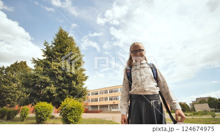 Cheerful schoolgirl with glasses and backpack standing near school, basking in sunlight under leafy trees and azure sky 124674975
