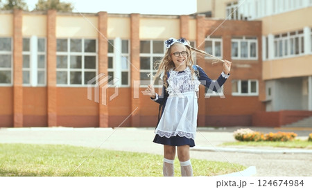 Cheerful schoolgirl twirling hair, standing near school entrance during bright, sunny afternoon 124674984