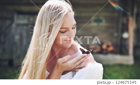Delicate blonde woman cradling butterfly on palm, serene natural setting with weathered wooden cabin behind her Delicate blonde woman cradling butterfly on palm, serene natural setting with weathered wooden cabin behind her 124675145