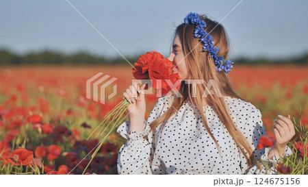 Blonde woman smiling, wearing blue flowers in hair, standing amid vibrant red poppy field and inhaling deeply near blooming blossoms 124675156