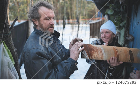 Impoverished couple gripping rusty pipe near weathered shelter during winter season Impoverished couple gripping rusty pipe near weathered shelter during winter season 124675292