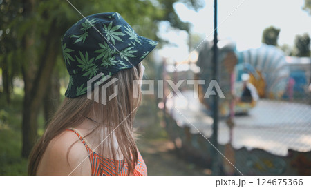 Female wearing cannabis themed headwear observing carnival ride amid sunlit park landscape 124675366