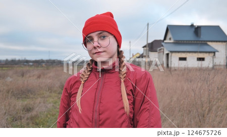 Portrait of a teenage girl wearing a red beanie and jacket, standing in a field with houses in the background Portrait of a teenage girl wearing a red beanie and jacket, standing in a field with houses in the background 124675726