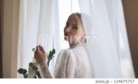 Elegant blonde bride wearing white gown, holding floral bouquet, gazing softly over shoulder near bright window Elegant blonde bride wearing white gown, holding floral bouquet, gazing softly over shoulder near bright window 124675757