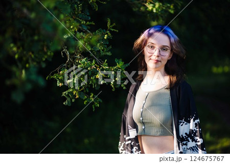 Stylish woman with purple hair and glasses leaning against tree, basking in warm summer sunlight amid green park backdrop 124675767