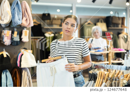 Young female buyer in store has removed hanger with trousers from window and is considering product. Young female buyer in store has removed hanger with trousers from window and is considering product. 124677648