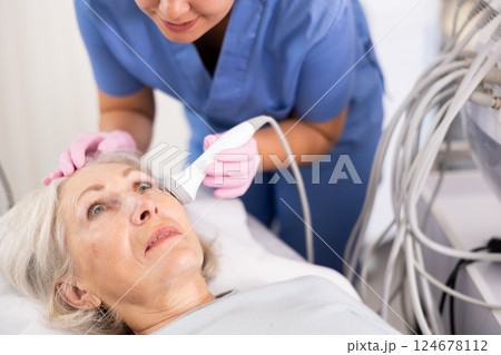 Young female doctor, dermatologist using ultrasound cavitation machine for improvement of skin condition of old female in clinic 124678112