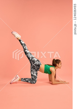 Young sporty girl wearing vibrant top and leggings, lifting leg high in donkey kick pose, focused on strength and flexibility, against soft peach background. 124686493
