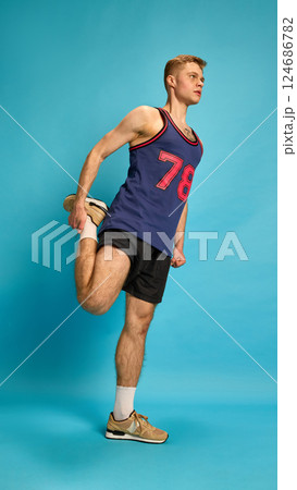 Athletic young man stretching leg back standing on one foot, showing flexibility and warm-up process, against studio background. Athletic young man stretching leg back standing on one foot, showing flexibility and warm-up process, against studio background. 124686782