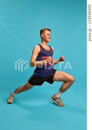 Sporty man doing side lunge with smile, dressed in shorts and tank top, showing strength and flexibility, against blue background. Sporty man doing side lunge with smile, dressed in shorts and tank top, showing strength and flexibility, against blue background. 124686800
