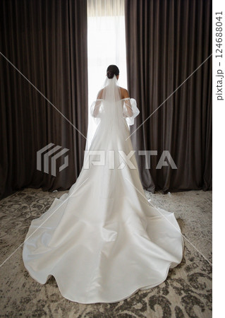 Bride wearing white wedding dress and veil looking out the window in hotel room Bride wearing white wedding dress and veil looking out the window in hotel room 124688041