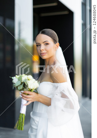 Bride walking near modern building wearing elegant white wedding dress and veil 124688045