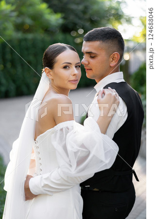 Groom tenderly embracing bride while holding hands in park during wedding ceremony 124688046