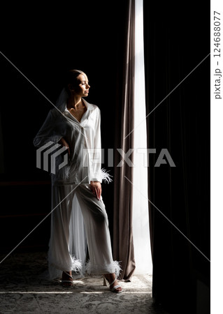 Bride opening curtains in white silk pajamas on wedding day 124688077