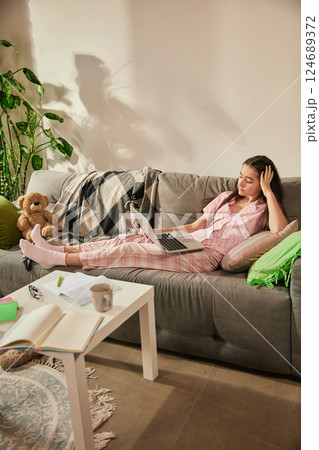 Teenage girl, student lying on couch with laptop, touching head in tiredness and boredom, attending online class, surrounded by notebooks and tea. 124689372