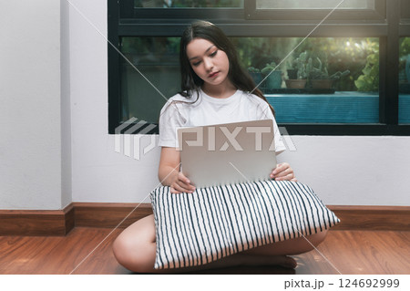 A young woman with laptop and pillow sitting drowsy on the living room floor. 124692999