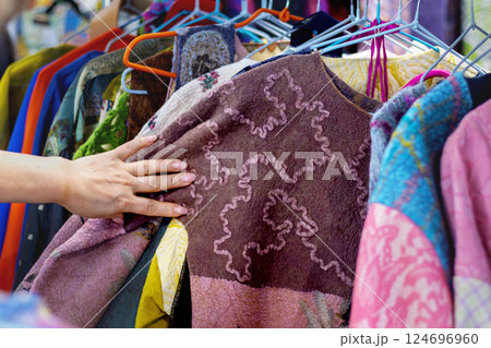 Woman examines brightly colored clothing at market, feeling textured felted wool fabric with voluminous embroidery showing unique patterns and variety of colors. Close-up of hand 124696960