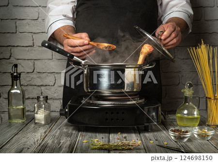 A chef prepares a simmering pot on a stove, adding spices and herbs while a wooden spoon stirs. The kitchen has a rustic vibe with wooden surfaces A chef prepares a simmering pot on a stove, adding spices and herbs while a wooden spoon stirs. The kitchen has a rustic vibe with wooden surfaces 124698150