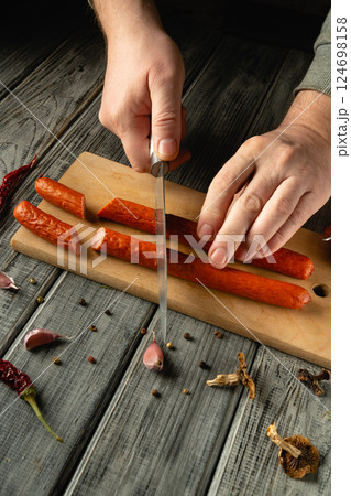 Hands expertly slice through savory sausages and garlic on a wooden cutting board. The kitchen has a warm atmosphere, enhanced by nearby spices and herbs 124698158