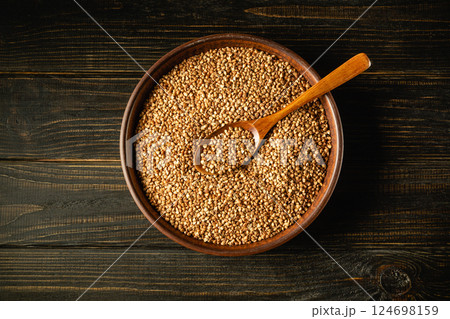 A wooden bowl filled with buckwheat grains sits on a dark wooden table. A wooden spoon sits among the buckwheat seeds, showing their texture and color in natural light. 124698159